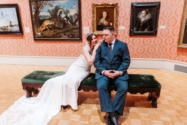 A bride in a white wedding dress and a groom in a blue suit sit on a green bench in an art gallery, with framed paintings on an orange patterned wall behind them.
