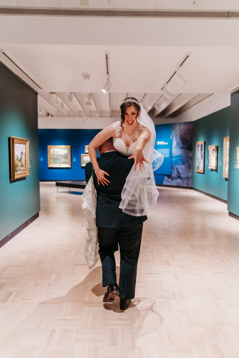 A man carries a woman in a white dress on his shoulders through a blue-walled art gallery.
