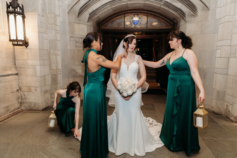 Bride in a white wedding gown holding a bouquet stands between two bridesmaids in emerald green dresses under a stone arch, while one kneels to adjust her train.
