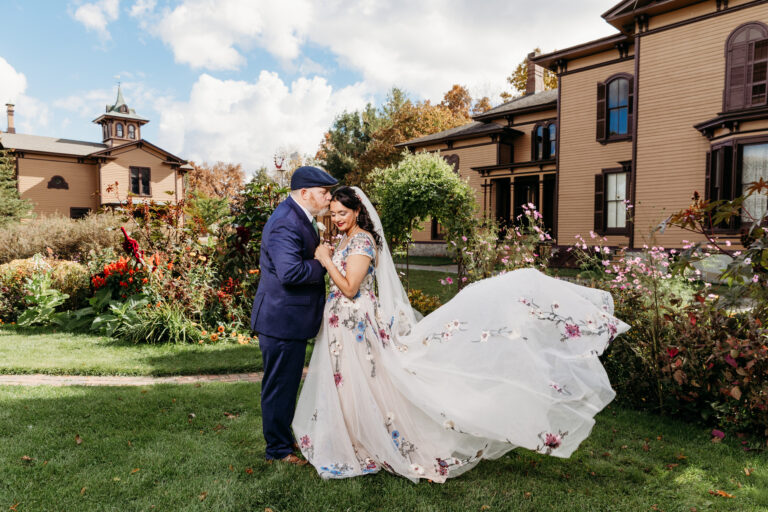 Bride and groom kiss in a garden with the bride's flowing white wedding dress and a brick villa in the background.