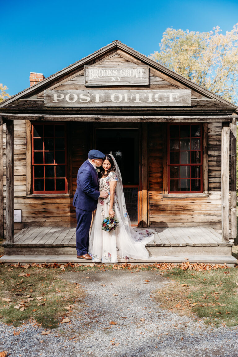 A bride and groom in wedding attire stand in the doorway of a rustic wooden post office on a sunny day.
