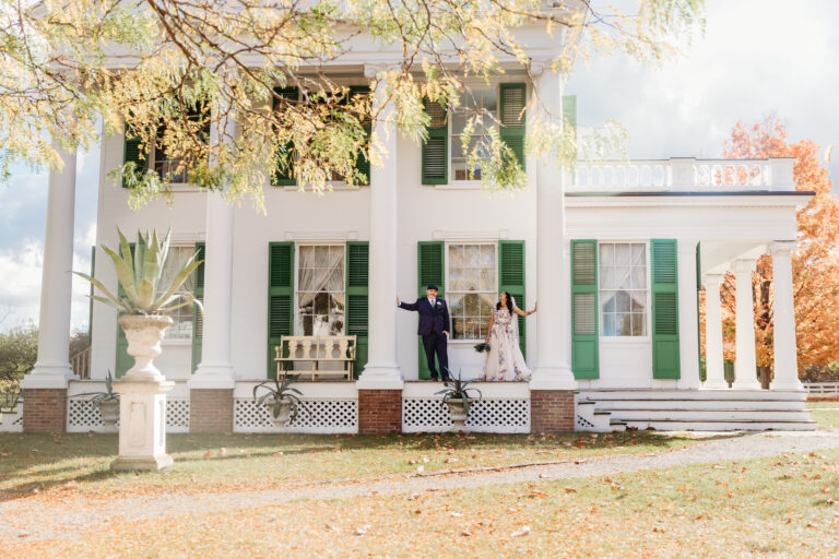 A white two-story house with a large front porch, green shutters, and two people standing on the steps amid fall leaves.