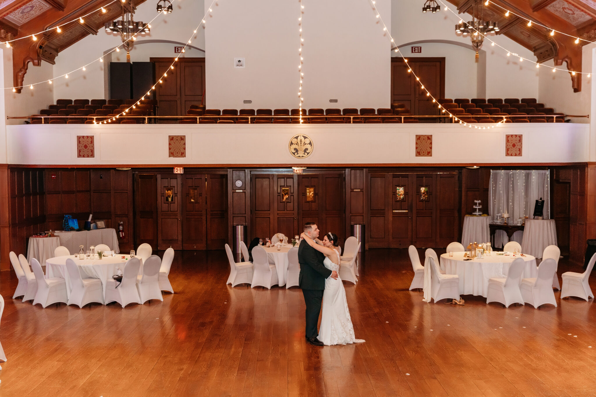 A couple dances in a spacious ballroom with a polished wooden floor, string lights overhead, and round tables dressed with white chair covers.