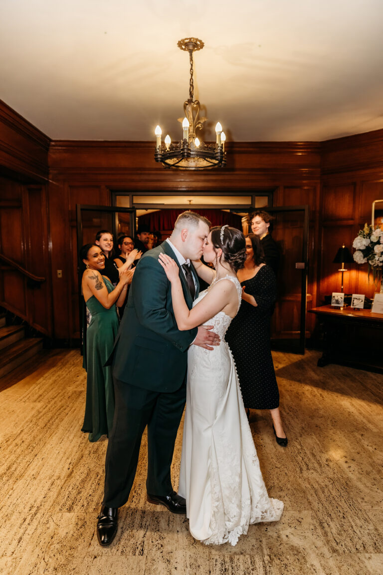 Bride and groom share a kiss in a wood-paneled room as formally dressed guests look on.