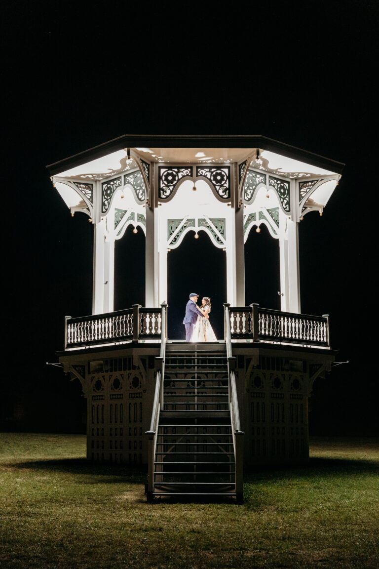 A couple in formal attire stands on the illuminated, ornate white gazebo at night.