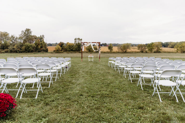 Outdoor wedding ceremony setup on a grassy field with white folding chairs arranged in rows facing a flower-decorated arch.