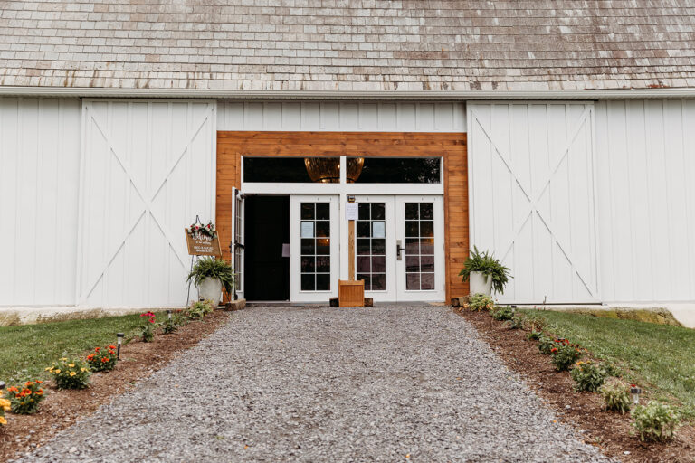 White barn-style building with a large wooden-framed glass doorway, flanked by potted plants and neatly lined flower beds along a gravel path.