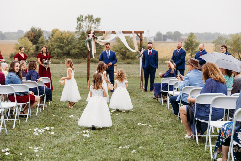 Outdoor wedding ceremony on a grassy field with a white wooden arch, flower girls in white dresses walking toward the altar while guests sit in white chairs.