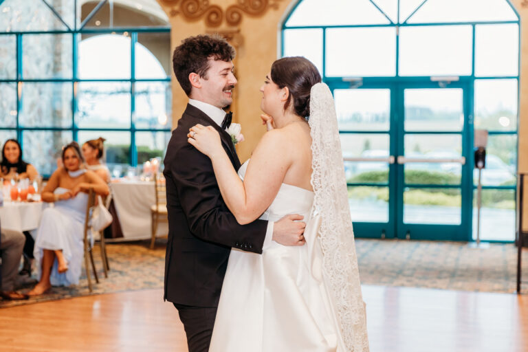 A bride and groom dance together at their wedding reception, with guests watching and clapping in the background.