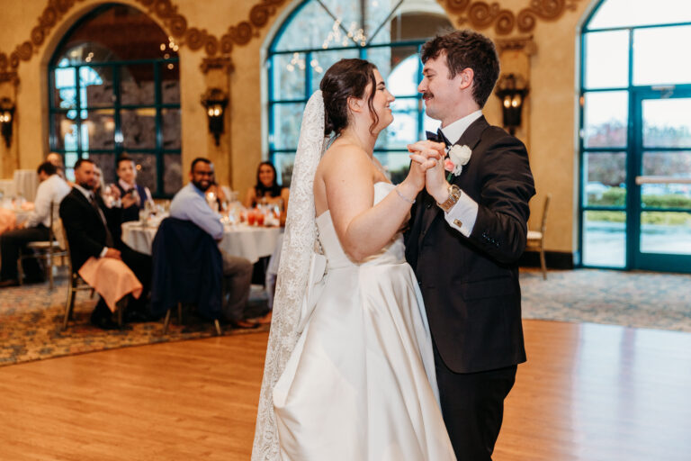 Bride and groom dancing together in a warmly lit wedding reception, with guests seated at tables in the background.