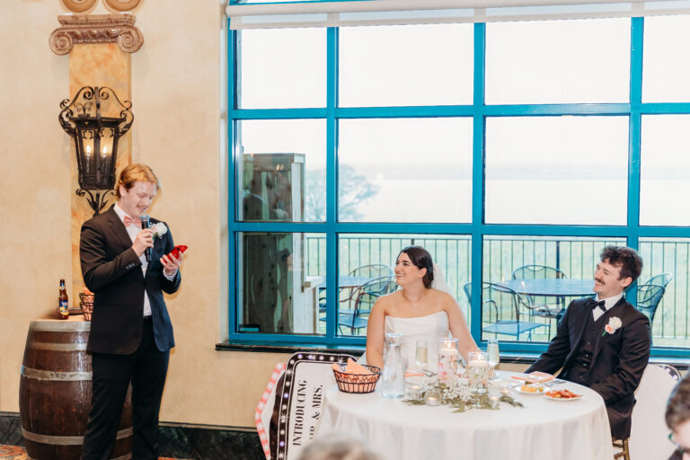 A bride and groom sit at a round table during a wedding reception as a man in a suit gives a toast beside a vintage barrel and lantern, with bright blue-framed windows in the background.
