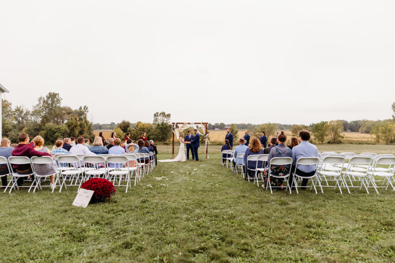 An outdoor wedding ceremony on a grassy field with white folding chairs set in rows along a central aisle and guests seated as a couple stands at the altar.