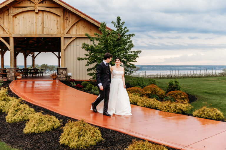 A bride in a white wedding dress and a groom in a dark suit walk along an orange curved walkway beside a wooden pavilion, with shrubs and a lake in the background.