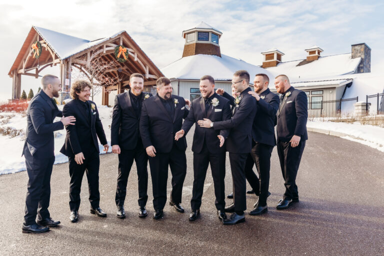 Group of men in black suits posing together outside a rustic wooden lodge in a snowy setting.