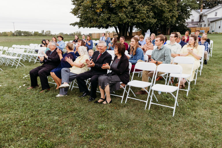 Group of guests seated on white folding chairs on a grassy lawn for an outdoor ceremony.