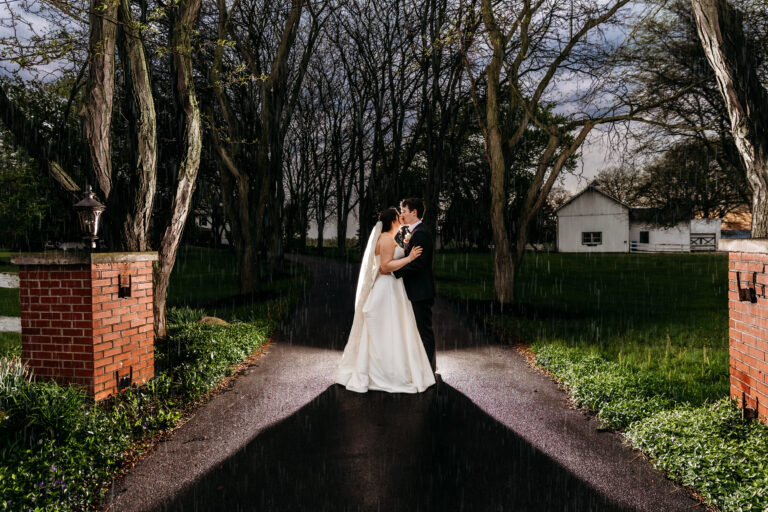 Bride and groom kiss on a tree-lined driveway framed by brick gate posts.