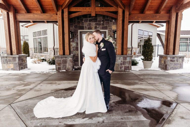 A bride and groom embrace under a wooden pavilion, the bride in a white wedding gown with a long train and the groom in a dark suit.