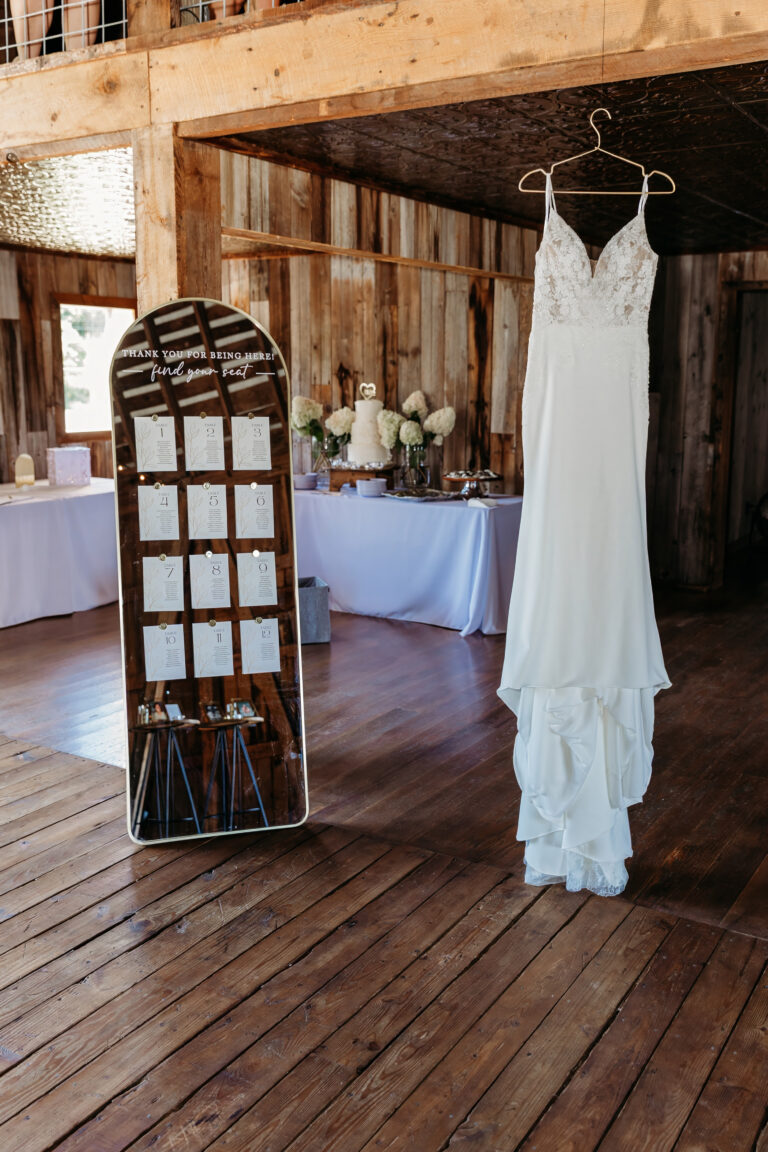 A white wedding dress hangs from a hanger in a rustic wooden hall with tables set for a reception in the background.