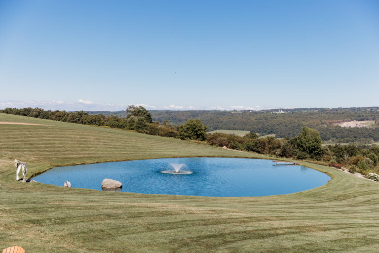A circular blue pool with a fountain sits in a grassy field under a clear blue sky, with rolling hills in the distance.