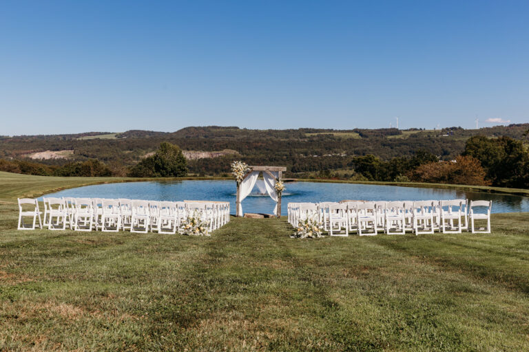 Outdoor wedding ceremony setup on a grassy field with white chairs arranged in rows along a central aisle and a white fabric arch at the end, with rolling hills and a blue sky in the background.