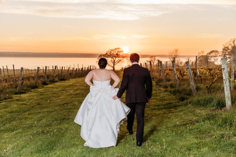 Bride in a white wedding dress and groom in a suit walk together along a grassy path between vineyard rows at sunset.