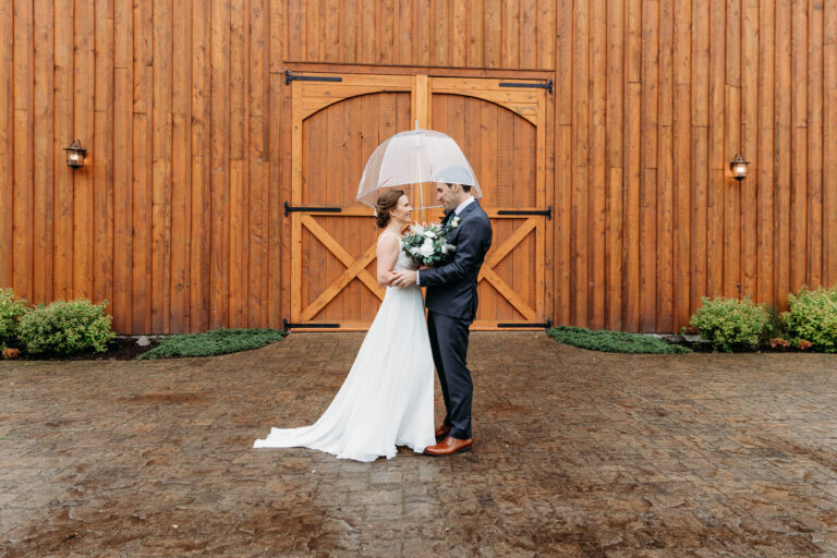 Bride and groom kiss under a white umbrella in front of a large wooden barn door.