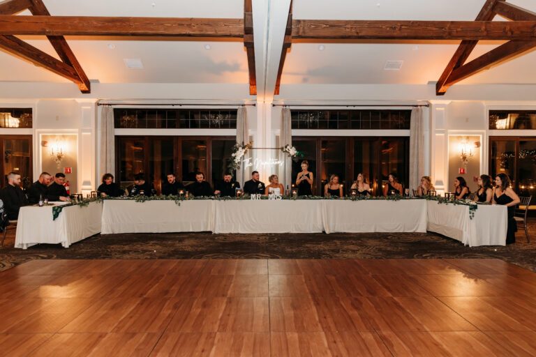 A long banquet table with guests seated on both sides in a warmly lit room with exposed wooden ceiling beams.