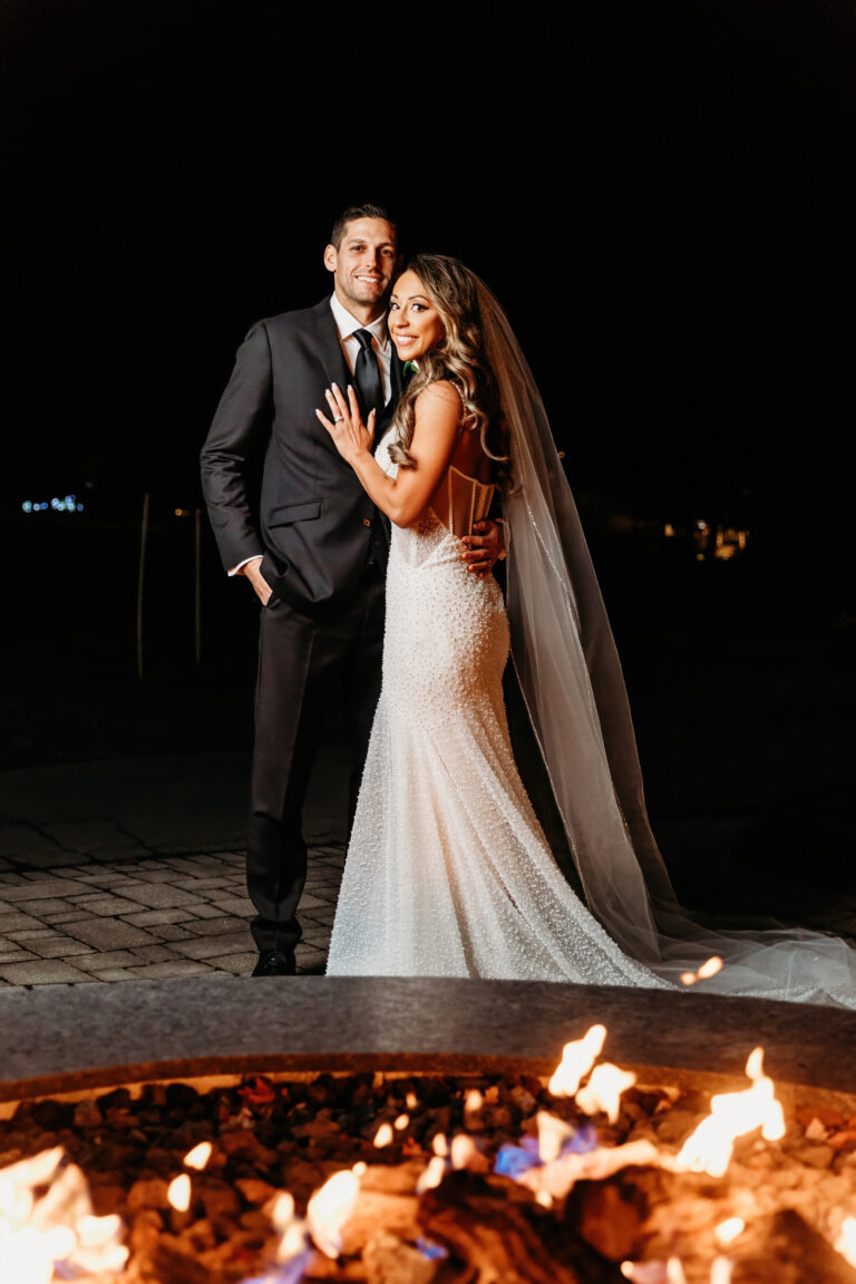 A bride in a white wedding gown with a long veil and a groom in a dark suit pose together by a lit fire pit at night.