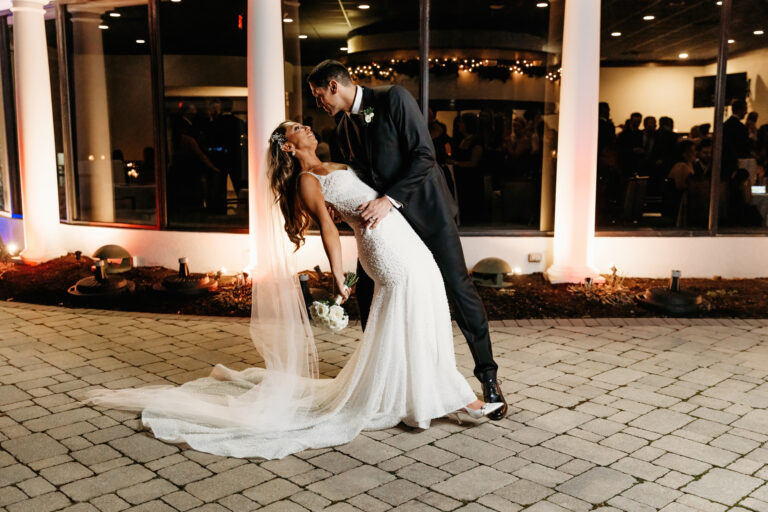 Bride and groom dip and kiss outdoors at night, the bride wearing a long white dress with a veil.