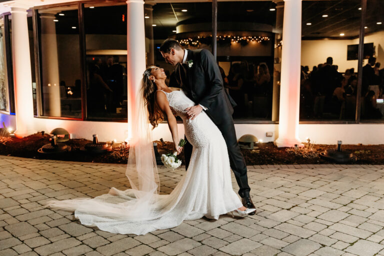 A bride in a white wedding dress and veil is being dipped by her groom outdoors at night, with string lights and a brick-paved ground in the background.
