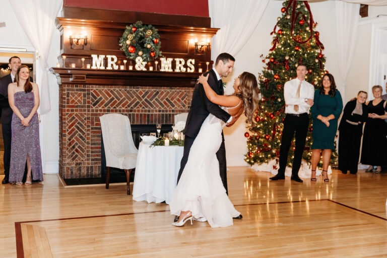 Bride and groom dancing at a wedding reception with a "MR & MRS" mantel, a Christmas tree, and guests watching.