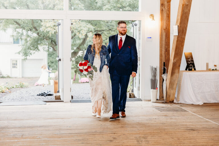 Bride and groom walk down a wooden indoor aisle, the bride in a white lace dress holding a red bouquet while the groom wears a navy suit with a burgundy tie.