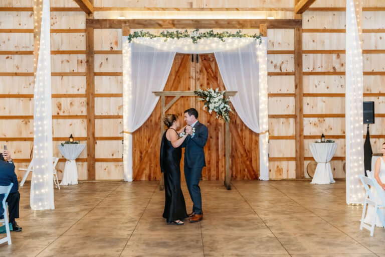 A couple dances in a rustic wooden barn wedding venue with a draped backdrop and greenery.