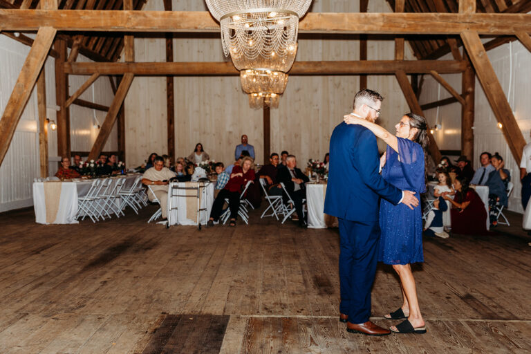 A couple dances in a rustic barn hall with a chandelier overhead, while guests sit at tables in the background.
