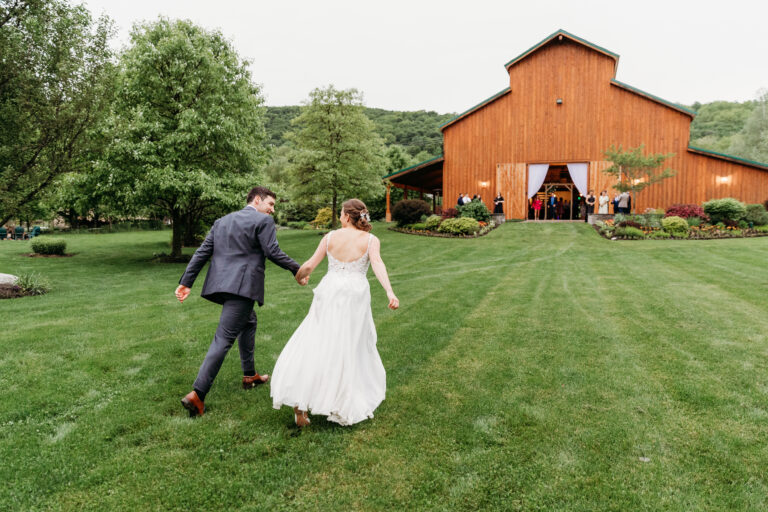 Bride and groom walk hand in hand across a grassy lawn toward a red barn at an outdoor wedding.