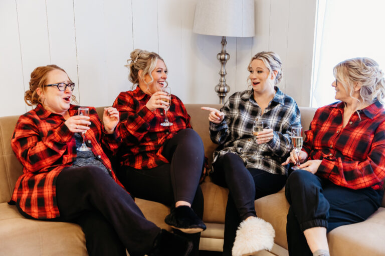 Four women in plaid shirts sit on a beige couch, drinking from mugs and chatting in a bright living room.