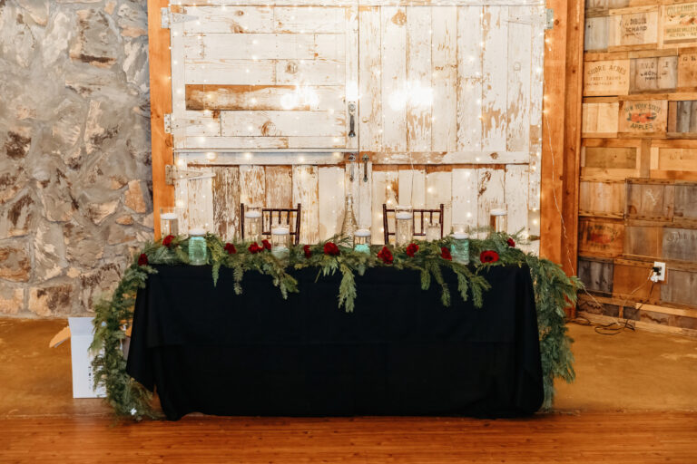 A long table covered with a black cloth is decorated with evergreen garlands and red accents in a rustic indoor setting.