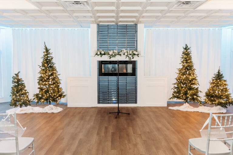 A bright, festive room decorated for Christmas with two illuminated trees on either side of a mantle and garland, and a microphone stand at the front.