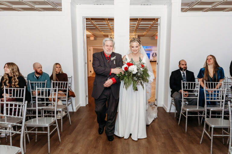 A bride in a white wedding gown carries a red-and-white bouquet while walking down the aisle with an older man in a black suit, as guests sit on gray chairs in a bright ceremony room.