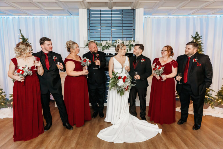 Centrally positioned bride in a white gown stands with bridesmaids in red dresses and groomsmen in black suits with red accents, posing indoors against white drapery.