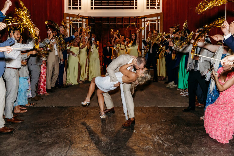 A couple performs a dance dip in the center of a wedding reception, while guests line up on both sides clapping.