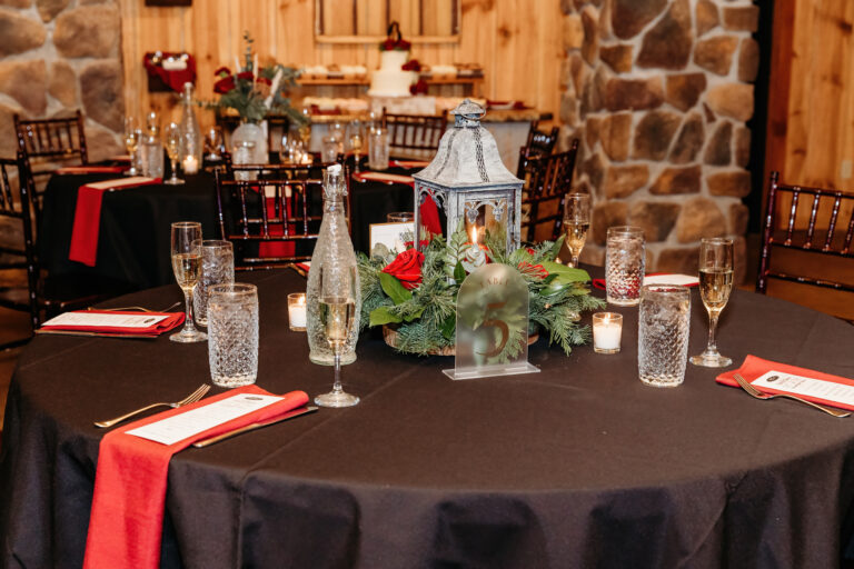 Round banquet table with a black cloth, red napkins, glassware, and a lantern centerpiece with greenery in a rustic wood-and-stone venue.