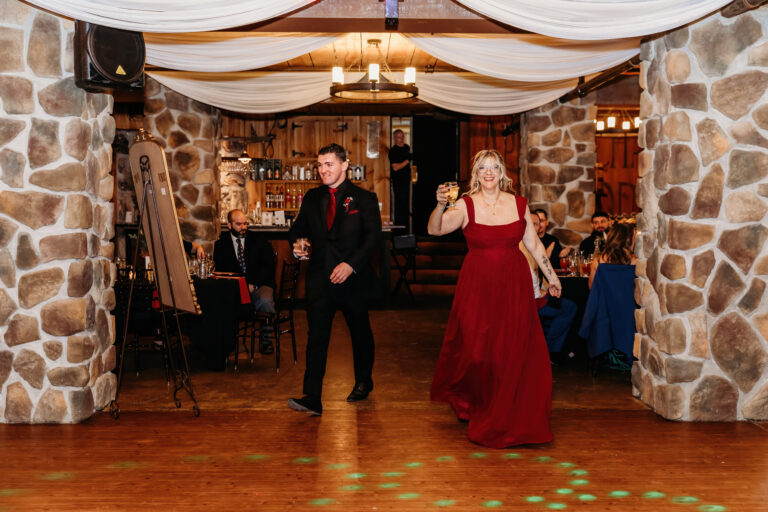A man in a black suit and a woman in a red dress walk across a wooden dance floor in a stone-walled event venue.