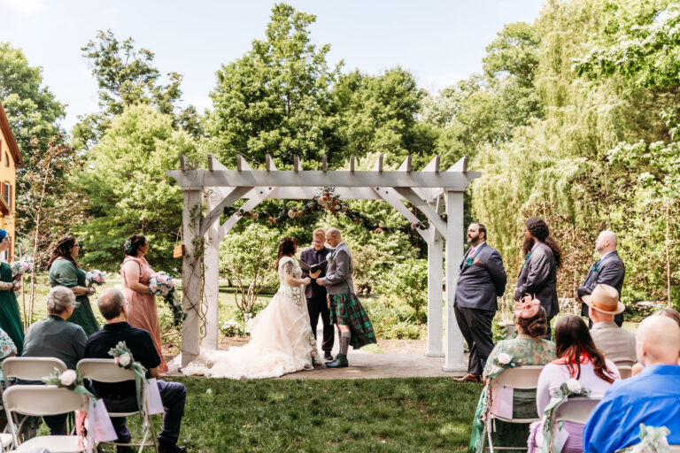 An outdoor wedding ceremony under a wooden arbor, with the bride and groom exchanging vows as guests sit on chairs along the aisle.