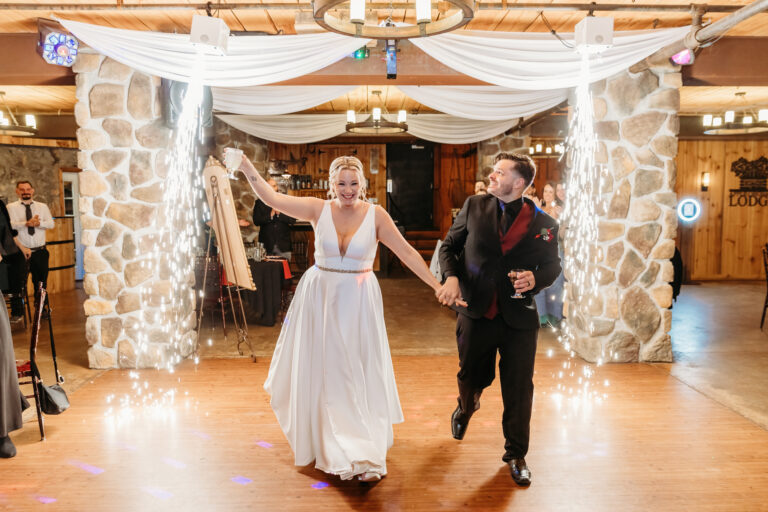 A bride in a white gown and a groom in a dark suit dance hand in hand beneath a draped stone arch at a rustic wedding reception.