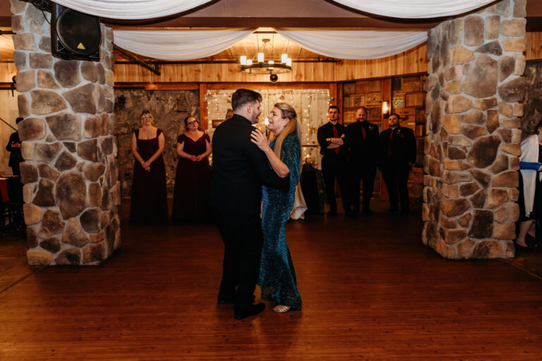 A couple dances closely on a wooden dance floor at a formal event, with guests and stone pillars visible in the background.