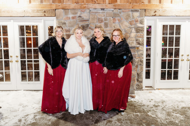 Bride in a white wedding dress poses with four bridesmaids in burgundy dresses and black jackets in front of a stone building with glass doors.