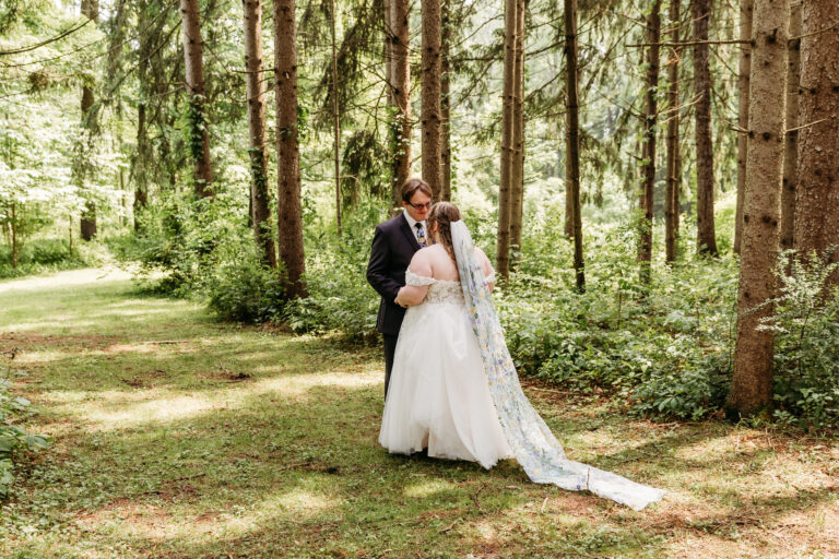 A bride and groom embrace in a sunlit forest, the bride in a long white wedding gown with a veil.
