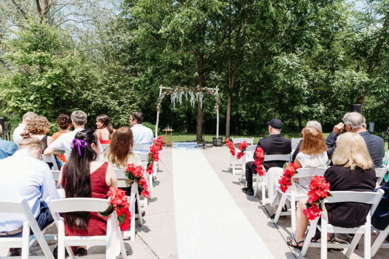 Outdoor wedding ceremony with guests seated on white chairs along a white aisle runner and a floral arch at the front.