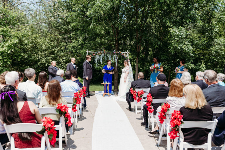 Outdoor wedding ceremony with a bride and groom at the altar under trees, guests seated along a white aisle lined with red flowers.